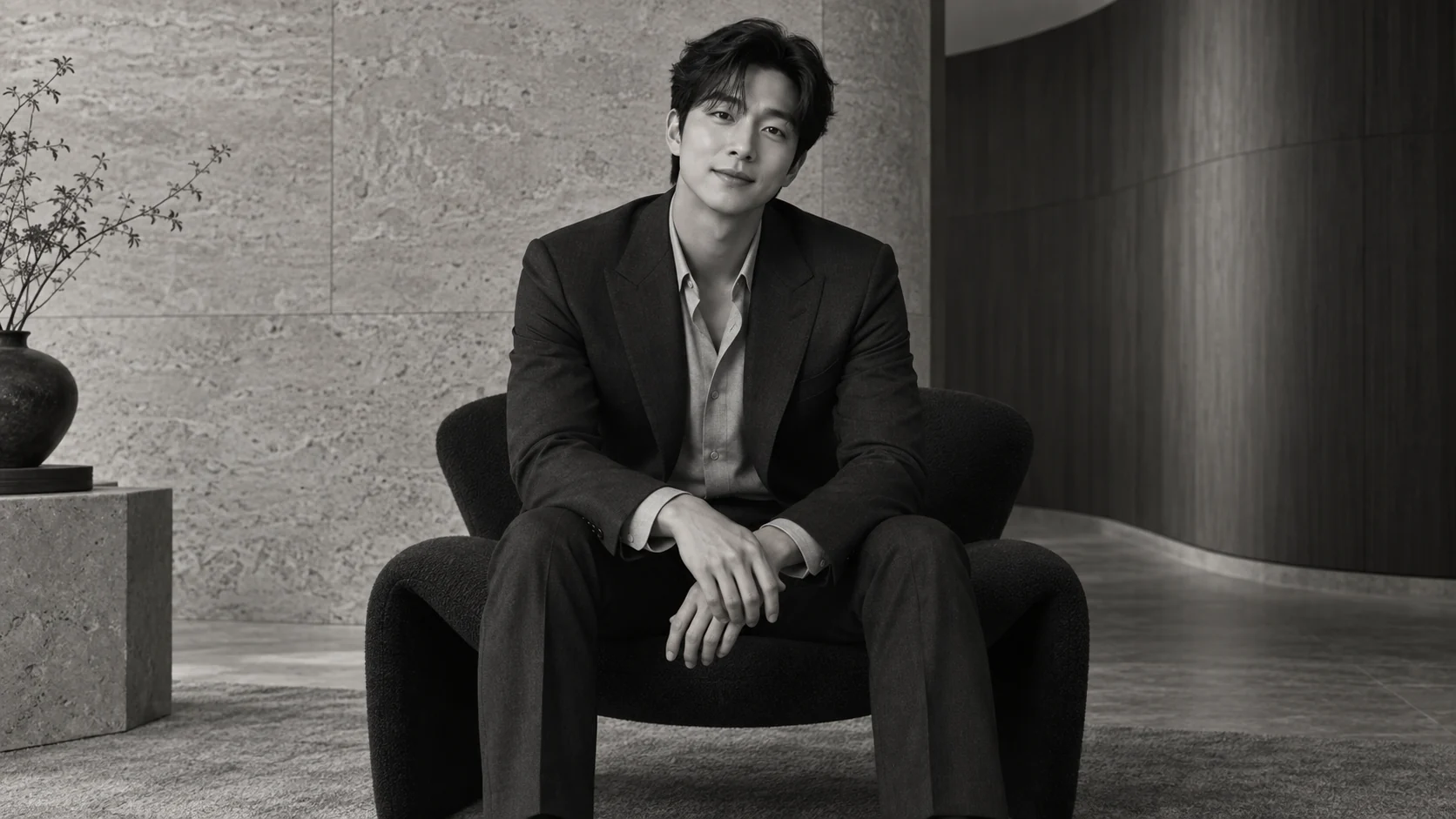 차콜 정장 차림의 남성이 어두운 의자에 앉아 있는 흑백 화보